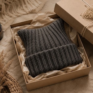 May include: A dark gray ribbed knit beanie with pointed cat-ear-like corners, presented inside a square cardboard gift box lined with crinkled tissue paper. The beanie features a thick folded cuff and a consistent vertical knit texture throughout.