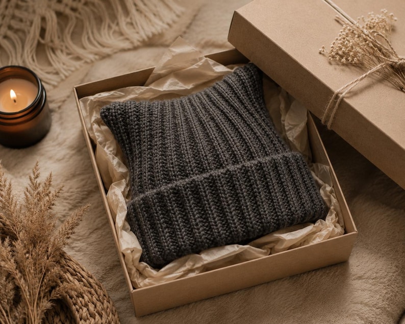 May include: A dark gray ribbed knit beanie with pointed cat-ear-like corners, presented inside a square cardboard gift box lined with crinkled tissue paper. The beanie features a thick folded cuff and a consistent vertical knit texture throughout.