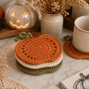 May include: A stack of three crocheted pumpkin-shaped coasters in orange, cream, and green. A matching orange coaster sits under a beige mug. A white pumpkin-shaped candle holder and a small orange pumpkin are in the background.