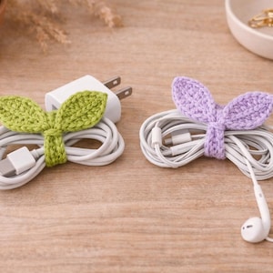 May include: Two cable organizers, one green and one purple, each shaped like leaves. The green organizer wraps around a white charging cable and plug. The purple organizer holds white earbuds. Both are on a wooden surface.