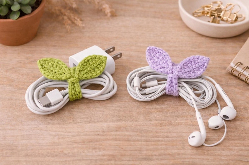 May include: Two cable organizers, one green and one purple, each shaped like leaves. The green organizer wraps around a white charging cable and plug. The purple organizer holds white earbuds. Both are on a wooden surface.