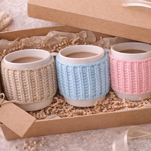 May include: A gift box containing three white mugs filled with a warm beverage. Each mug is adorned with a knitted cozy in beige, light blue, and pink. The box is lined with paper and includes a blank tag.