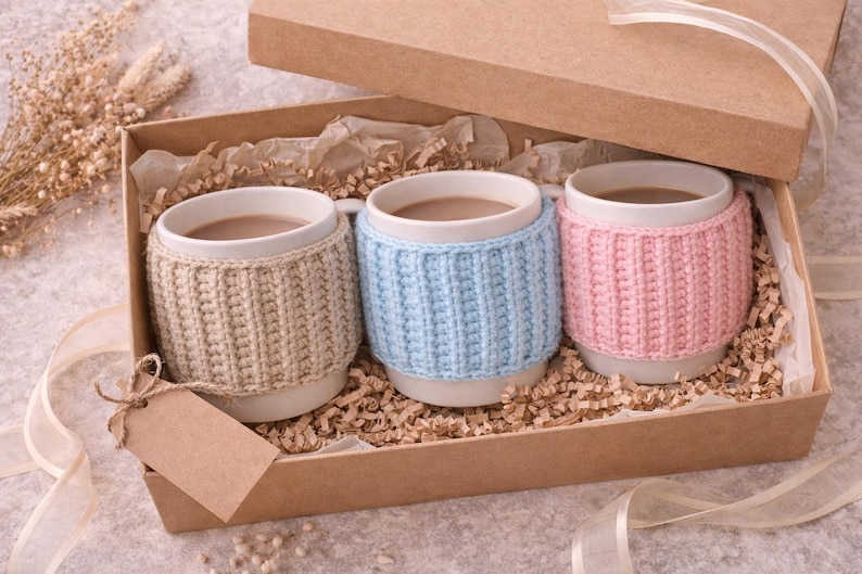 May include: A gift box containing three white mugs filled with a warm beverage. Each mug is adorned with a knitted cozy in beige, light blue, and pink. The box is lined with paper and includes a blank tag.