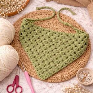 May include: A handmade, green crochet bandana with a square pattern, displayed on a woven mat. The bandana has tie strings. The image also shows white yarn balls, pink scissors, a crochet hook, and decorative elements.