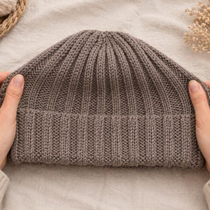 May include: A ribbed knit beanie in a neutral taupe color. The hat is held by two hands, showcasing the texture and shape of the warm winter accessory. The beanie is made of a soft, knitted material.