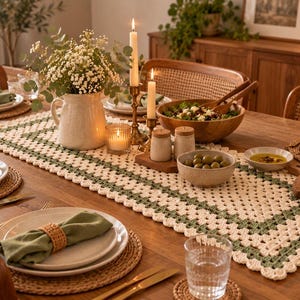 Op de afbeelding: Een rechthoekige gehaakte tafelloper met een crèmekleurig midden en een saliegroene rand, geplaatst op een houten eettafel met witte borden, saliegroene servetten, gevlochten placemats, een keramieken kan en kaarsen.