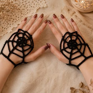 May include: A pair of black fingerless gloves featuring a crocheted spider web pattern. The gloves cover the back of the hands and are secured with a loop around the middle finger, leaving the fingers and palms exposed for full dexterity.