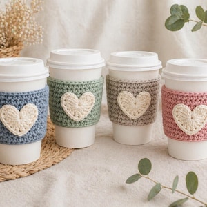 May include: Four white paper coffee cups with lids, each adorned with a crocheted cozy. The cozies are in blue, green, gray, and pink, each featuring a cream-colored heart. The cups sit on a woven mat, with greenery and a vase in the background.