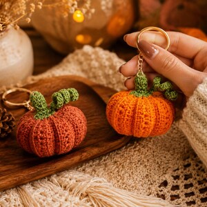 Puede incluir: Dos llaveros de calabaza de ganchillo. Uno est&aacute; sostenido por una mano, el otro descansa sobre una bandeja de madera. Las calabazas son naranjas con tallos verdes. El fondo incluye un jarr&oacute;n decorativo, una pi&ntilde;a y una superficie de punto color crema.