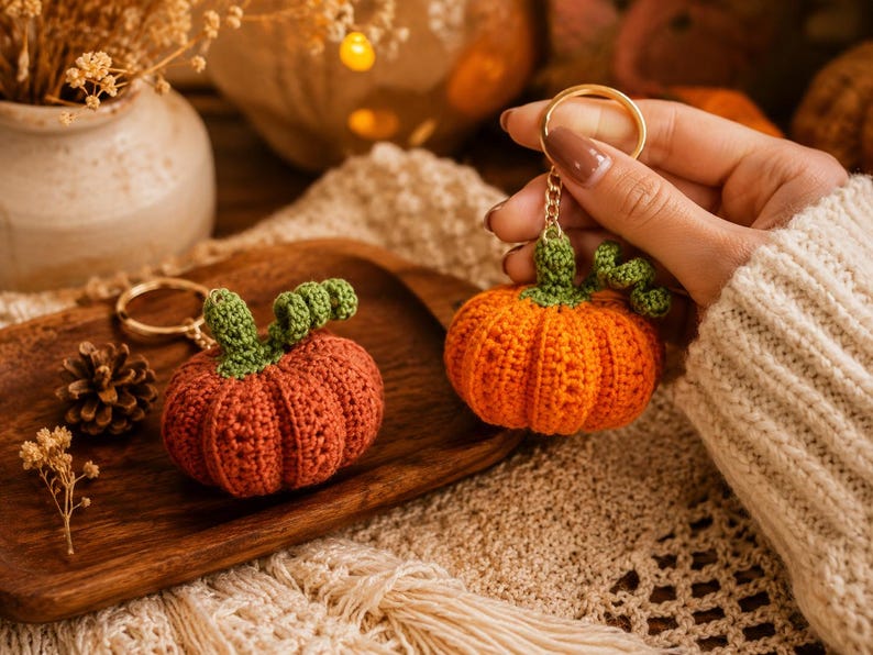 Puede incluir: Dos llaveros de calabaza de ganchillo. Uno est&aacute; sostenido por una mano, el otro descansa sobre una bandeja de madera. Las calabazas son naranjas con tallos verdes. El fondo incluye un jarr&oacute;n decorativo, una pi&ntilde;a y una superficie de punto color crema.