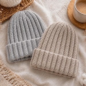 Könnte beinhalten: Zwei gerippte Strickmützen in Hellgrau und Beige liegen auf einer weichen, weißen Oberfläche. Die Mützen haben einen umgeschlagenen Rand und ein klassisches, schlichtes Design. Die Szene wird durch eine Tasse Kaffee und dekorative Elemente ergänzt.
