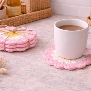 May include: A white mug filled with coffee sits on a pink and white crochet coaster. A stack of matching coasters tied with twine is nearby. The coasters are daisy-shaped, with pink petals and a yellow center. The scene suggests a cozy kitchen setting.