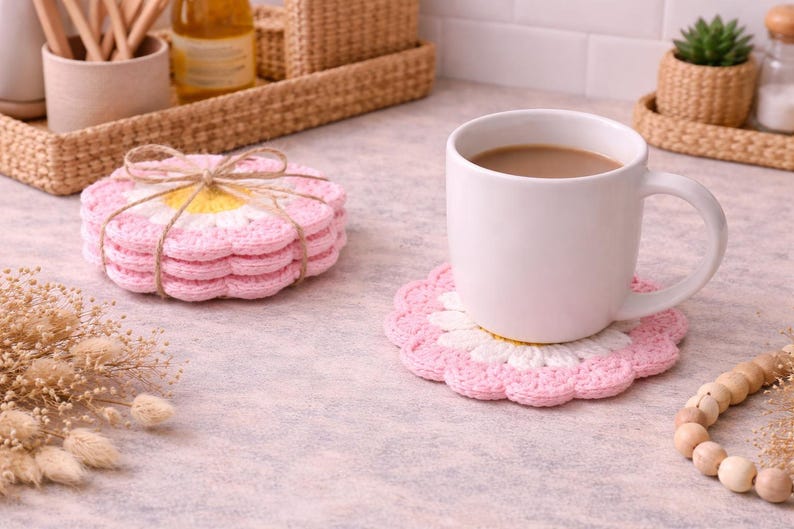 May include: A white mug filled with coffee sits on a pink and white crochet coaster. A stack of matching coasters tied with twine is nearby. The coasters are daisy-shaped, with pink petals and a yellow center. The scene suggests a cozy kitchen setting.