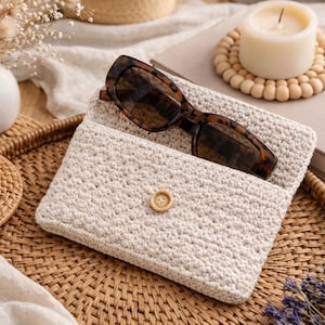May include: A cream-colored, crocheted sunglasses case with a wooden button closure. Tortoiseshell sunglasses rest inside the open case. The case sits on a woven tray with a cup of coffee, a candle, and a book.