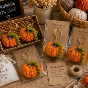 Puede incluir: Llaveros de calabaza naranja de ganchillo con enredaderas verdes y llaveros dorados. Los llaveros est&aacute;n empaquetados en tarjetas marrones que dicen "handmade with love". Una peque&ntilde;a bolsa dice "gracias".