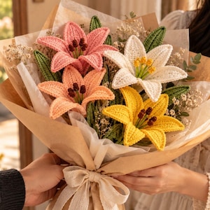 May include: A bouquet of four crocheted lily flowers in pink, peach, white, and yellow, each featuring dark brown stamens and green crocheted leaves. The flowers are wrapped in brown paper and white tissue paper, tied with a beige ribbon.