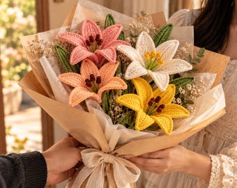 May include: A bouquet of four crocheted lily flowers in pink, peach, white, and yellow, each featuring dark brown stamens and green crocheted leaves. The flowers are wrapped in brown paper and white tissue paper, tied with a beige ribbon.