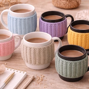 May include: A collection of ceramic mugs filled with a warm beverage, each adorned with a knitted cozy in various pastel colors. The mugs are arranged on a wooden surface, alongside cookies and a notepad.