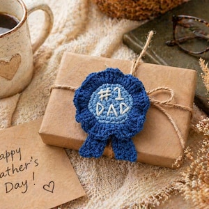 May include: A wrapped gift with a blue crocheted ribbon that says "#1 Dad." A coffee mug with a heart design and a card that says "Happy Father's Day!" are also in the image. The background is a neutral color.