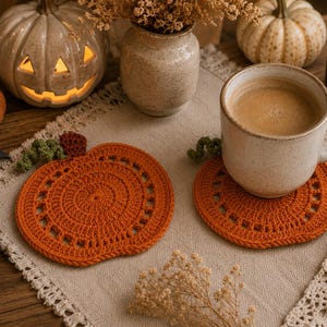 May include: Two orange crochet pumpkin coasters with a cup of coffee on one. The coasters are on a beige placemat with a pumpkin and a lit candle in the background. The scene evokes a cozy autumn atmosphere.