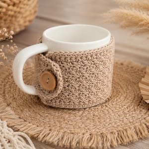 May include: A white ceramic mug featuring a removable beige crochet cozy. The cozy is crafted with a textured stitch pattern and is secured around the mug with a single round wooden button located near the handle, providing a snug fit.
