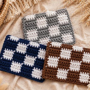 May include: Three handmade crochet coasters in a checkerboard pattern. One is navy blue and white, another is brown and white, and the third is gray and white. The coasters are square and rest on a beige surface.