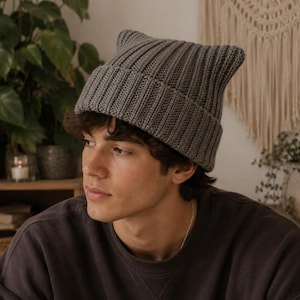 May include: A charcoal gray knit beanie featuring a ribbed texture and two pointed ears at the top. The hat has a wide, folded cuff that sits above the forehead, constructed from a thick, uniform yarn in a solid, neutral dark gray color.
