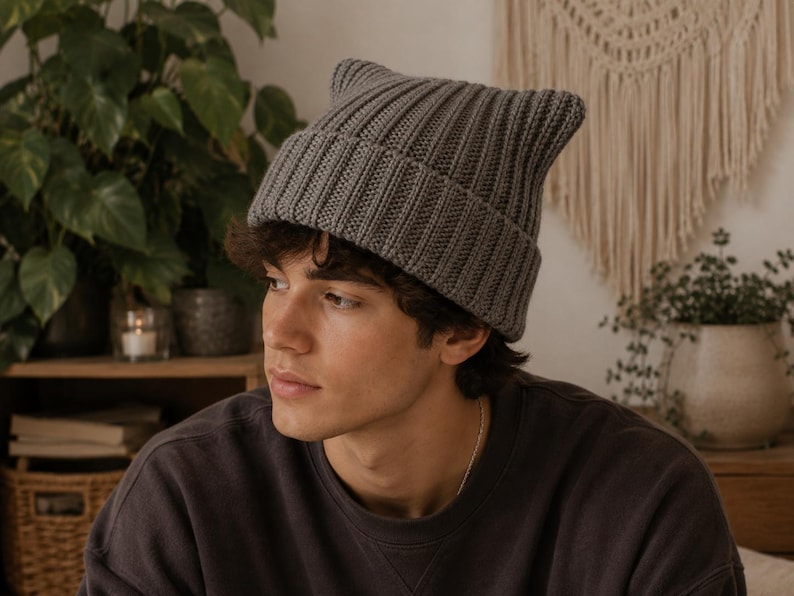 May include: A charcoal gray knit beanie featuring a ribbed texture and two pointed ears at the top. The hat has a wide, folded cuff that sits above the forehead, constructed from a thick, uniform yarn in a solid, neutral dark gray color.