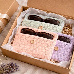 May include: A gift box containing three pairs of sunglasses in crocheted cases. The cases are pastel colors: light green, peach, and lavender. Each case has a wooden button closure. A separate gift box with a ribbon is also present.