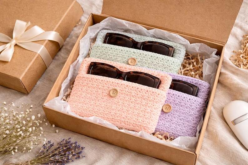 May include: A gift box containing three pairs of sunglasses in crocheted cases. The cases are pastel colors: light green, peach, and lavender. Each case has a wooden button closure. A separate gift box with a ribbon is also present.