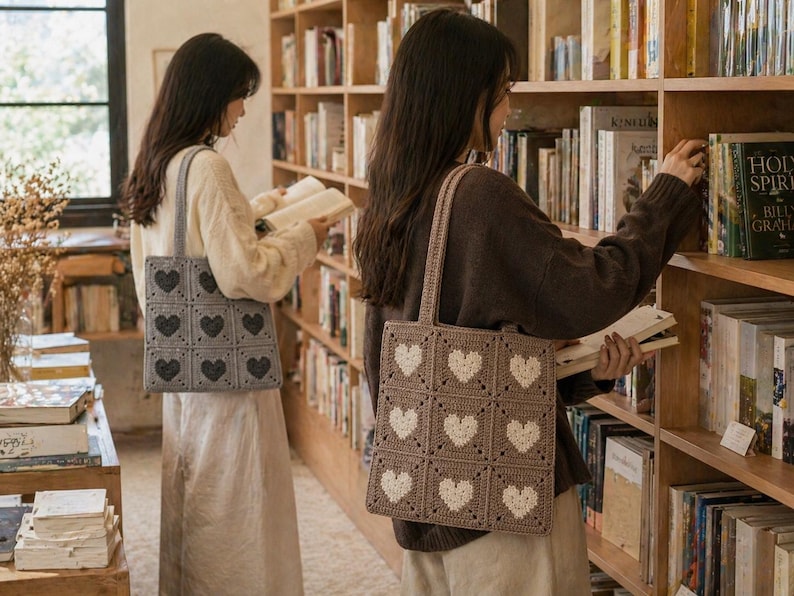 May include: Two women in a library each carry a crocheted tote bag. One bag is gray with dark gray hearts, the other is tan with white hearts. The bags are square and have a handle. The women are browsing bookshelves filled with books.