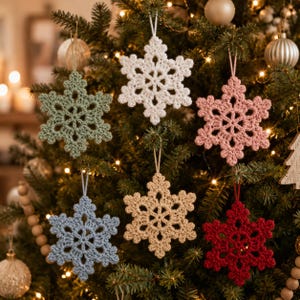 May include: A collection of five crocheted snowflake ornaments hanging on a Christmas tree. The snowflakes are in white, sage green, dusty pink, light blue, and deep red, each featuring a six-pointed star design with intricate yarn loops.