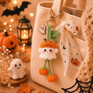 May include: A handmade Halloween-themed keychain featuring a crocheted ghost wearing a pumpkin hat. The keychain is attached to a beige tote bag with a ribbon adorned with pumpkins and stars. Decorative pumpkins, a bat, and a lantern are in the background.