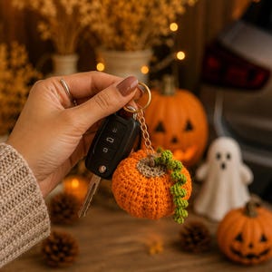 Puede incluir: Un llavero de calabaza naranja tejido a ganchillo con detalles de vid verde, sujeto a un mando de coche. El fondo est&aacute; borroso, con decoraciones de Halloween, incluyendo calabazas y un fantasma.