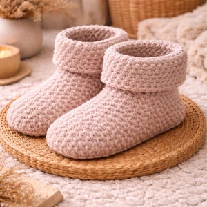 Op de afbeelding: Een paar lichtroze, handgehaakte pantoffels met een omgeslagen manchet. De pantoffels staan op een ronde, geweven mat. Deze gezellige pantoffels zijn ontworpen voor gebruik binnenshuis en bieden warmte en comfort.