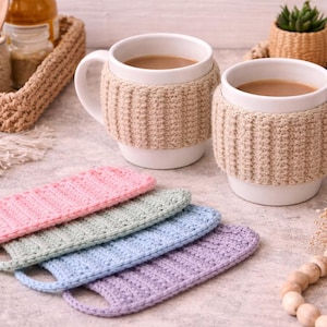 Puede incluir: Dos tazas blancas con café, cada una envuelta en una funda de punto beige. Cuatro posavasos de punto en colores pastel, rosa, verde, azul y morado, están en primer plano. Una escena acogedora y hogareña.