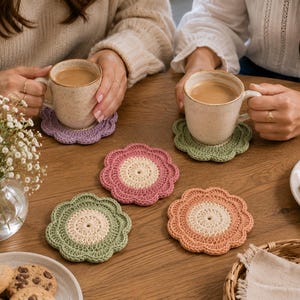 Puede incluir: Un conjunto de cinco posavasos de ganchillo con forma de flor en varios colores, incluyendo lavanda, verde salvia, rosa empolvado y melocotón. Cada posavasos presenta un centro de color crema sólido con un borde festoneado.