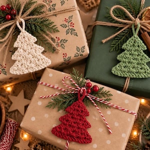 May include: Three gift boxes wrapped in brown and green paper, each decorated with a crocheted Christmas tree ornament in cream, green, or red. The packages feature twine, evergreen sprigs, red berries, dried orange slices, and cinnamon sticks.