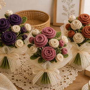 May include: Three crochet flower bouquets in purple, pink, and orange. Each bouquet is wrapped in white tulle and tied with a cream-colored ribbon. The flowers are arranged with green leaves and cream-colored buds.