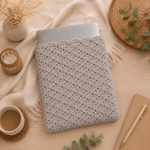 May include: A light gray laptop sleeve featuring a textured, star-like crochet pattern with a scalloped top edge. The sleeve is constructed from yarn and is designed to hold a flat, rectangular electronic device securely within its soft, woven structure.