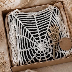 May include: A white crochet top with a spiderweb design, presented in a brown cardboard box with dried flowers and a blank tag. The top is displayed on a black surface, creating a striking contrast.