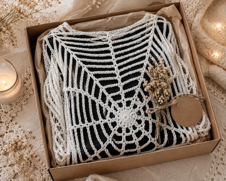 May include: A white crochet top with a spiderweb design, presented in a brown cardboard box with dried flowers and a blank tag. The top is displayed on a black surface, creating a striking contrast.