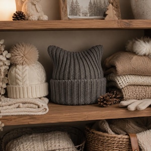May include: A dark gray knit beanie with pointed cat-ear details and a wide ribbed cuff. The beanie is positioned on a wooden shelf alongside a cream-colored cable knit beanie with a large pom-pom, folded sweaters, and knit gloves.