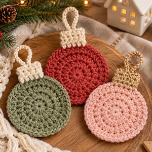 May include: Three circular crochet ornaments in olive green, deep red, and soft pink. Each ornament features a textured, round body and a small, rectangular hanging loop at the top. The green and red ornaments have cream-coloured loops, while the pink ornament has a metallic gold loop.