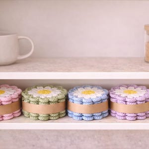 May include: A shelf displays five crochet daisy-shaped boxes in pastel colors: pink, green, blue, purple, and cream. Each box has a yellow center and a brown band. A white mug and a jar with a wooden lid are also on the shelf.