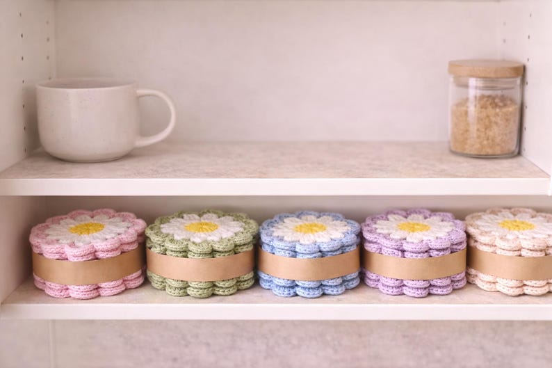 May include: A shelf displays five crochet daisy-shaped boxes in pastel colors: pink, green, blue, purple, and cream. Each box has a yellow center and a brown band. A white mug and a jar with a wooden lid are also on the shelf.