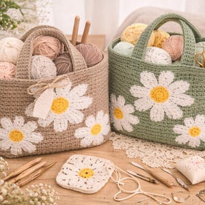 May include: Two crochet yarn storage baskets, one tan and one green, each adorned with white daisy appliqués with yellow centers. The baskets are filled with yarn balls in various colors. Knitting supplies and a cup of coffee are also visible.