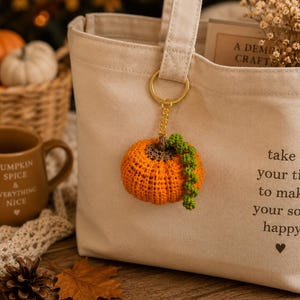 Puede incluir: Un llavero de calabaza naranja de ganchillo con enredaderas verdes cuelga de un bolso tote beige. La bolsa tiene el texto "take your time to make your soul happy". Una taza marr&oacute;n con texto est&aacute; cerca de una cesta de calabazas.