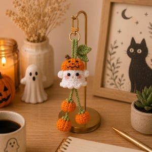 May include: A handmade Halloween-themed keychain featuring a crocheted ghost wearing a pumpkin hat. The ghost has a white body, orange pumpkin hat, and three dangling orange pumpkins. The keychain is attached to a gold-colored metal clasp.