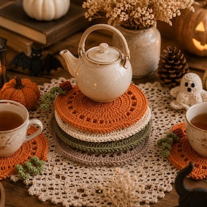May include: A cozy autumn-themed still life featuring a stack of crocheted orange, cream, green, and brown coasters. A teapot sits atop the coasters, with two cups of tea, pumpkins, and decorative items. The scene evokes a warm, inviting atmosphere.